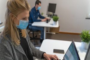 A woman in the foreground and a man in the background work on their computers, socially distanced and wearing masks